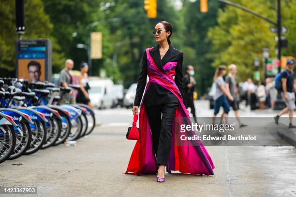Chriselle Lim wears black circle sunglasses, a black blazer jacket with red / purple / fuchsia long train detail, black suit pants, purple plastic...