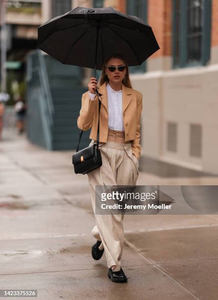 Fashion Week Guest seen wearing a beige cardigan, a white shirt, beige pants, a black leather handbag and a black umbrella and black sunglasses...