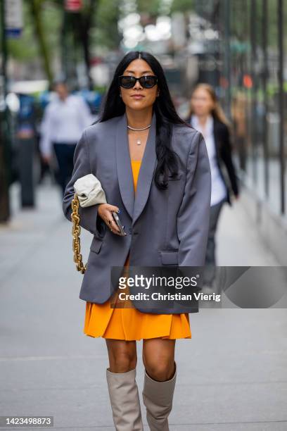 Guest wearing grey oversized blazer, orange dress, white Bottega Veneta bag, knee high boots outside Brandon Maxwell on September 13, 2022 in New...