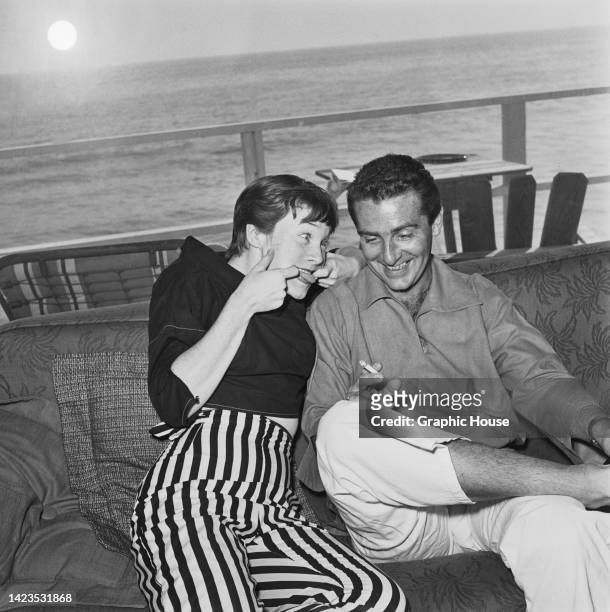American actress and singer Shirley MacLaine, wearing a black sweater and striped trousers, poking her tongue out at her husband, American...
