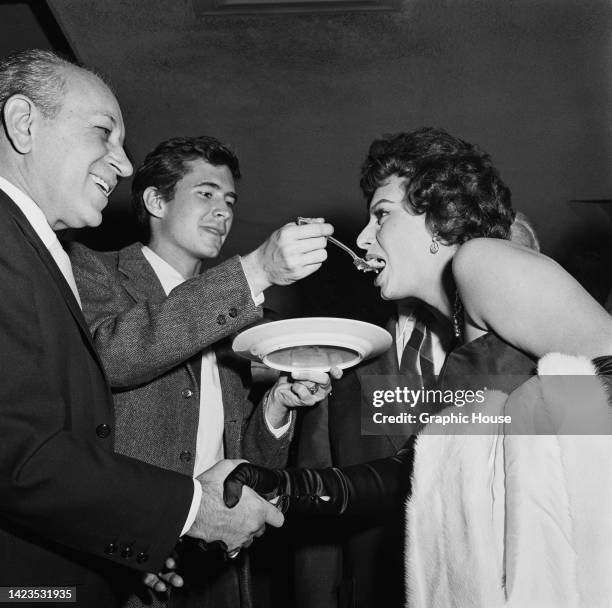 Low angle view of American actor George Raft shaking hands with Italian actress Sophia Loren who is being fed by American actor Anthony Perkins who...