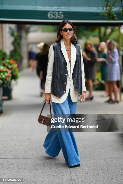 Jen Azoulay wears black sunglasses, gold necklace, a white latte blazer jacket, a black shiny leather sleeveless gilet, blue faded denim wide legs...