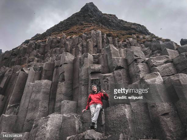 low angle view happy teenage boy sitting high up on basalt columns at reynisfjara black sand beach - black sand iceland stock pictures, royalty-free photos & images