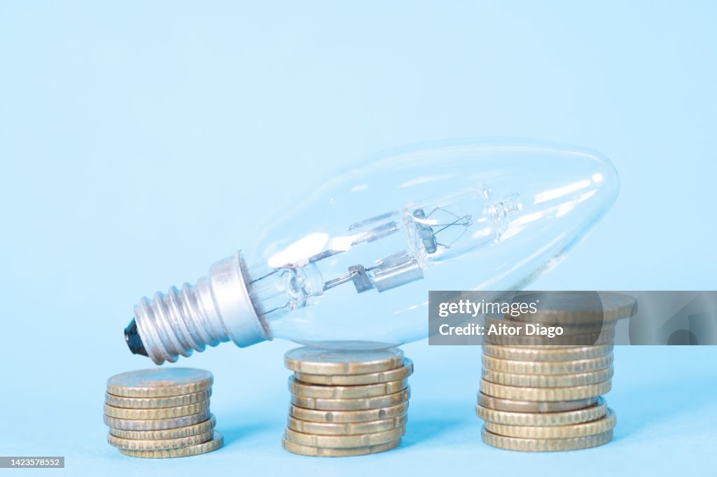 Picture representing the increase in electricity bills in times of inflation. Light bulb on coins three piles of coins in order from smallest to largest quantity.