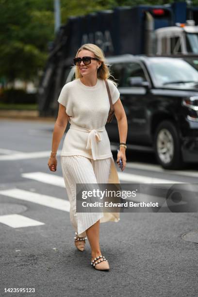 Guest wears black square sunglasses, a beige belted wool t-shirt, white latte pleated / accordion midi skirt, gold and silver chain bracelet, a gold...