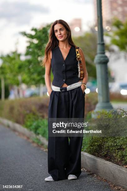 Gala Gonzalez wears a gold chain necklace, a black striped print pattern V-neck / buttoned / sleeveless gilet, black large pants, a beige and brown...