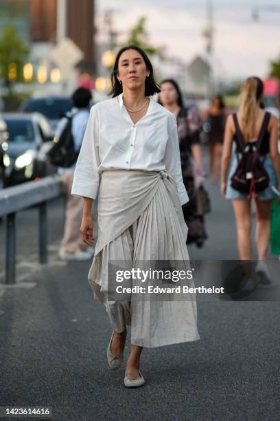 Eva Chen wears gold earrings, a gold necklace, a white shirt, pale gray plated / accordion wrap long skirt, gray shiny leather ballerinas , outside...