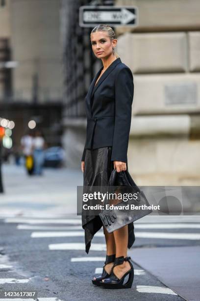 Xenia Adonts wears black sunglasses, silver and diamonds flash of lightning pendant earrings, a black blazer jacket, a black shiny leather shoulder...