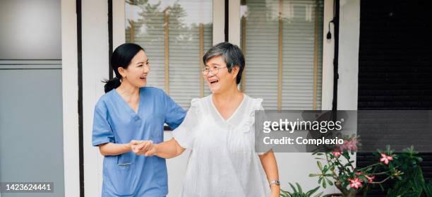 infirmière optimiste en uniforme bleu soutenant une femme âgée asiatique tout en marchant ensemble à travers la porte à l’extérieur de la maison - elderly care japanese photos et images de collection