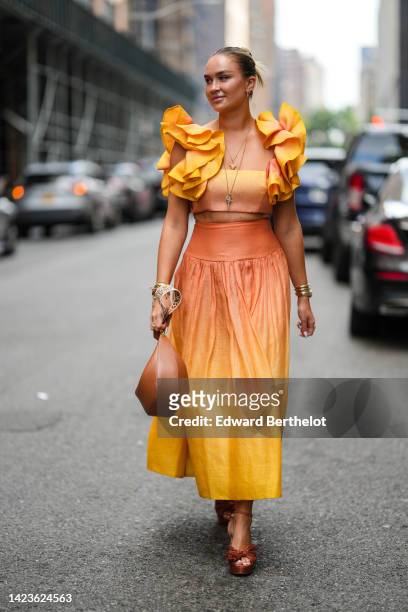 Nina Suess wears earrings, gold long chain necklaces, a yellow and orange gradient print pattern square neck / ruffled short sleeves cropped top, a...