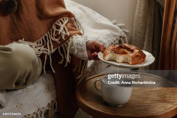 a woman holds a plate with a bitten bagel with poppy seeds on her bed. homemade dessert and cup of tea. wooden background. vintage scandi style. close-up - woman eating bagel stock pictures, royalty-free photos & images
