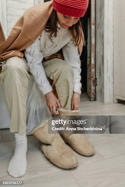 a woman in a sweater and beige pants puts on warm ugg boots. close-up. it's cold in the house. life in conditions of saving heat energy. - sheepskin boot fotografías e imágenes de stock