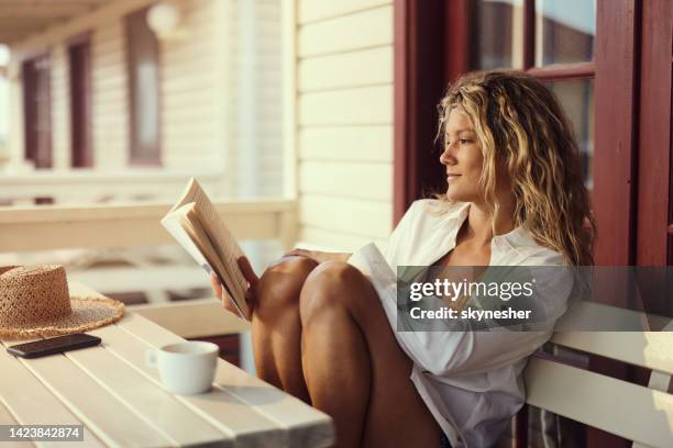 young relaxed woman reading a book on a bench at patio. - escapism reading stock pictures, royalty-free photos & images