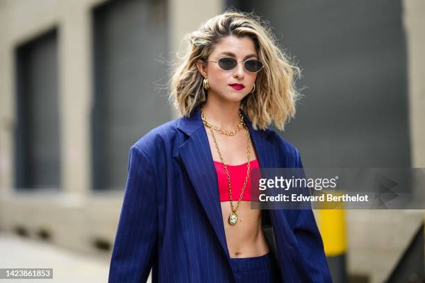 Guest wears black sunglasses, gold large earrings, a gold long chain necklace, a red V-neck / cropped top, a navy blue striped print pattern...