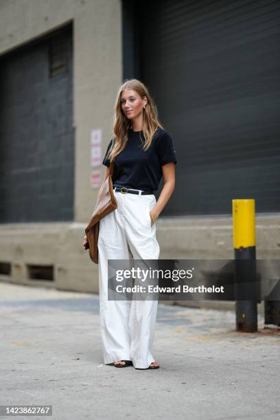 Guest wears gold earrings, a black t-shirt, a black shiny leather belt, white wide legs pants, a brown shiny oversized shoulder bag, flip flop heels...