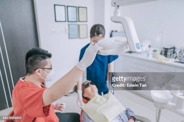 アジアの中国人歯科医は、彼の女性看護師の助けを借りて、高齢の女性患者のための歯科手術の準備を外科用ライトを調整する - 歯痛 ストックフォトと画像