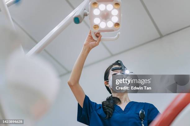 asiatische chinesische zahnärztin mit lupe, die das chirurgische licht von oben auf den patienten richtet - zahnarztstuhl stock-fotos und bilder