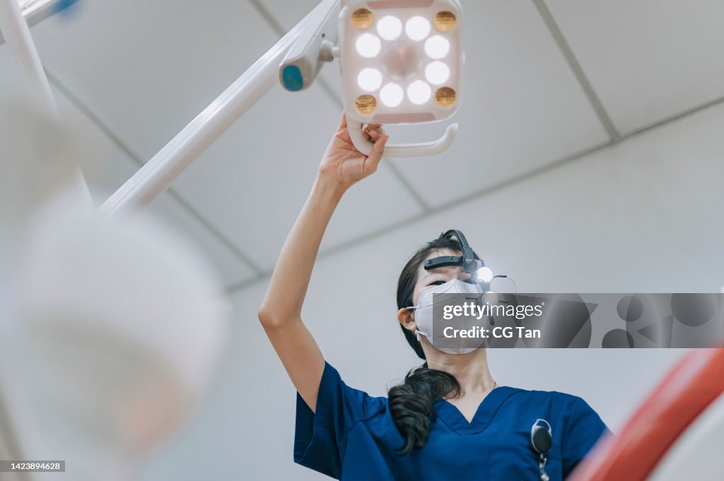 Asian chinese female dentist with magnifying glasses adjusting surgical light looking from above at patient