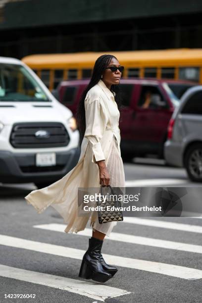 Guest wears black sunglasses, gold earrings, a white latte shirt / long pleated dress, a black and beige jacquard print pattern handbag from Prada,...