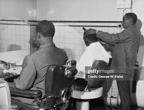 Jamaican barber George Clifford working on the hair of an unspecified man as another waits his turn, in a barbershop, location unspecified, borough...