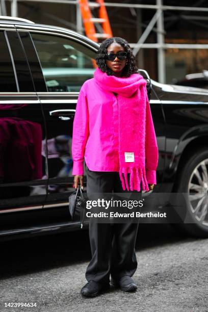 Guest wears black sunglasses, a neon pink oversized wool fringed scarf from Cos, a neon pink shirt, black large pants, a black nylon handbag, black...