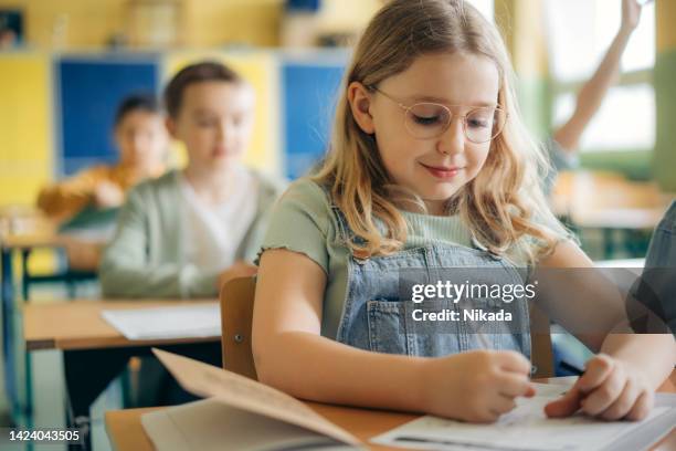 porträt eines süßen grundschulmädchens, das im klassenzimmer am schreibtisch sitzt - kind brille stock-fotos und bilder