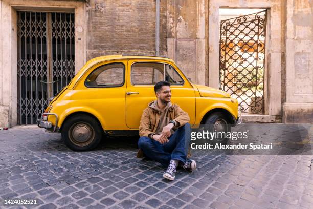 happy smiling man sitting next to a small yellow car in rome, italy - funny tourist stock pictures, royalty-free photos & images