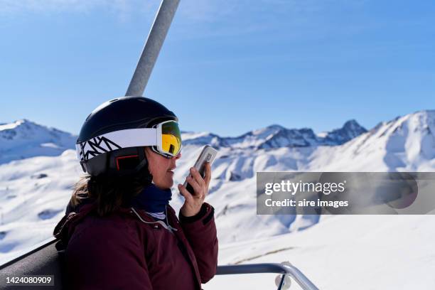 older female adult on a chairlift being carried while using her phone, on a clear, sunny day, wearing ski protection gear - winter sport - fotografias e filmes do acervo