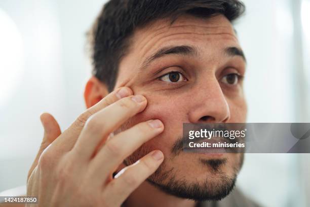 portrait of a handsome man applying cream under his eyes in the bathroom - man eye cream stock pictures, royalty-free photos & images