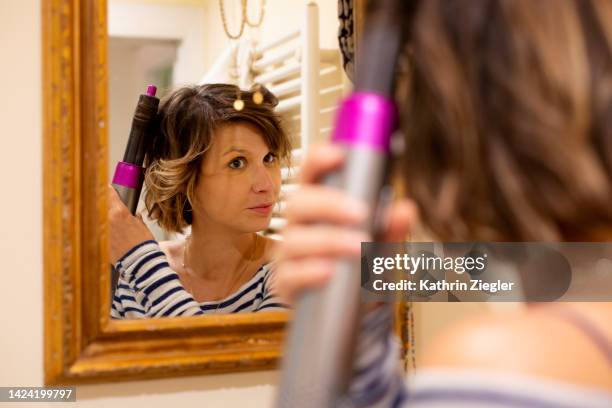 woman curling her hair in front of bathroom mirror - lockenstab stock-fotos und bilder