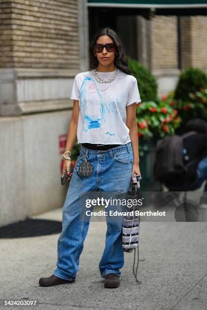 Guest wears black sunglasses, silver large chain necklaces, a white with pale blue and pink painted pattern t-shirt, blue denim large pants, a black...