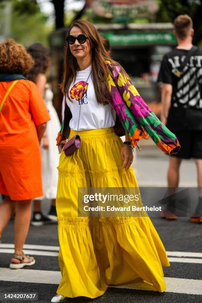 Guest wears black sunglasses, a white with red and yellow print pattern t-shirt, a purple with yellow / green / pink flower print pattern jacket, a...