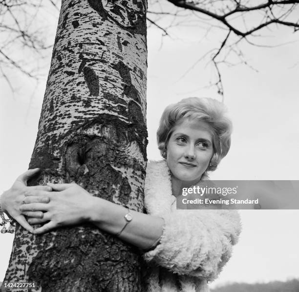 British film actress Liz Fraser , wearing a fleece coat, her arms around the trunk of tree, United Kingdom, 17th March 1961.