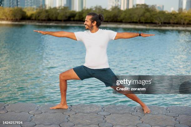 man practicing yoga by the sea in abu dhabi, standing in warrior 2 pose - warrior position stock pictures, royalty-free photos & images
