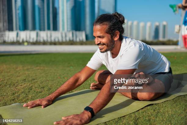 happy man doing yoga on lawn in public park in abu dhabi, sitting in easy pose and bending forward - yoga office arab stock pictures, royalty-free photos & images