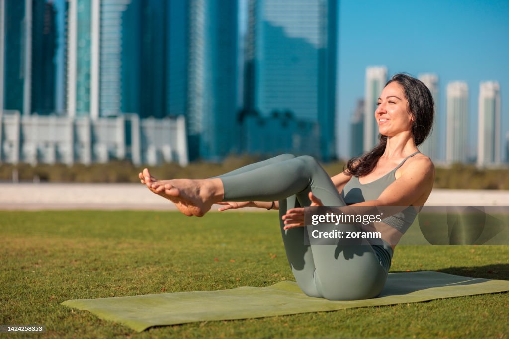 Woman doing yoga on lawn in public park in Abu Dhabi, sitting in Boat pose