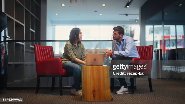 asian businesswoman and caucasian businessman sitting on sofa and working together on laptop in modern office working space - 2 men chatting casual office stock pictures, royalty-free photos & images