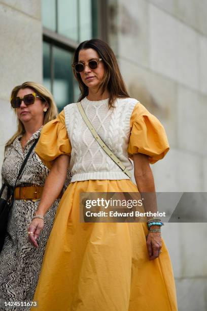 Guest wears brown sunglasses, an orange puffy short sleeves / long dress, a white latte braided wool sleeveless pullover, a white latte leather...