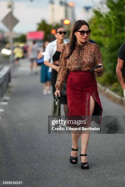 Guest wears black sunglasses, a brown / khaki / red print pattern shirt, a red suede / studded borders / split knees skirt, a khaki matte leather...