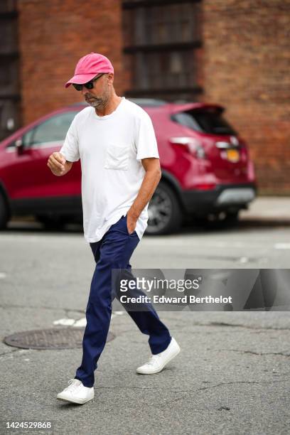 Guest wears a pink denim cap, black sunglasses, a white t-shirt, navy blue suit pants, white shiny leather sneakers , outside Gabriela Hearst, during...