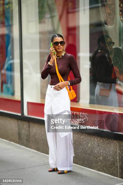 Guest wears black sunglasses, gold earrings, a gold chain necklace, a brown long sleeves t-shirt, an orange shiny leather crossbody bag, white cargo...