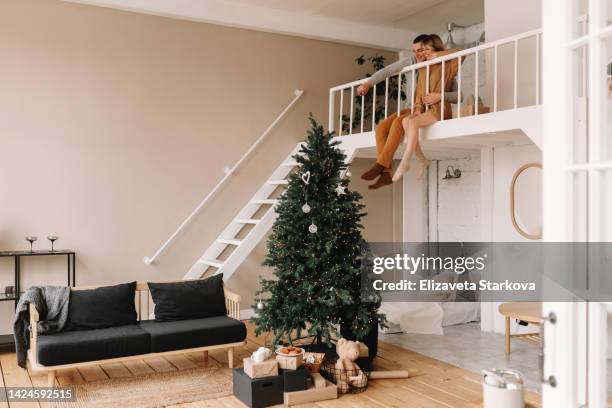 a happy couple in love in pajamas and red sweaters spend time together preparing for the christmas holiday, giving each other gift boxes in the new year sitting on the sofa in a cozy decorated room with a wooden floor, stairs and a christmas tree - christmas tree stockfoto's en -beelden