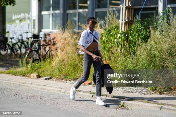 Guest wears brown sunglasses, a white with embroidered black pattern short sleeves shirt, a black crossbody bag, black denim skinny pants, a black...