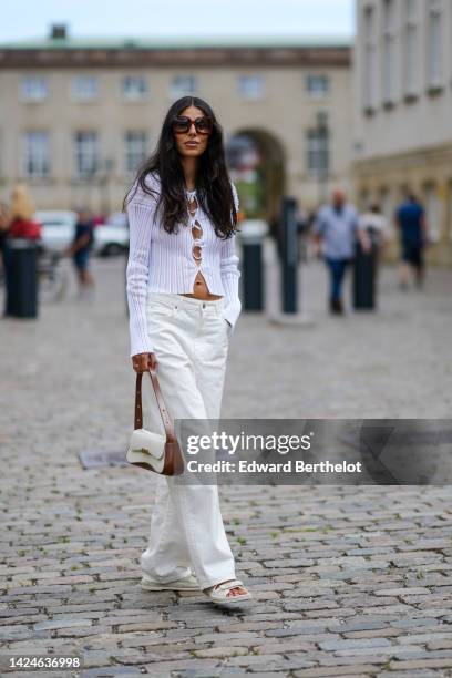 Guest wears brown sunglasses, a white ribbed wool / laces front cardigan, white denim large pants, a white leather and brown borders handbag, white...