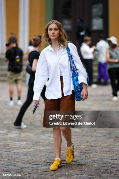 Guest wears a white shirt, brown long shorts, a blue print pattern oversized shoulder bag, white socks, yellow shiny leather with black logo pattern...