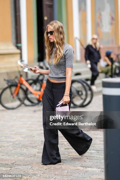 Guest wears black sunglasses, a gray short sleeves t-shirt, black striped print pattern wide legs print pattern pants, a pale purple matte leather...