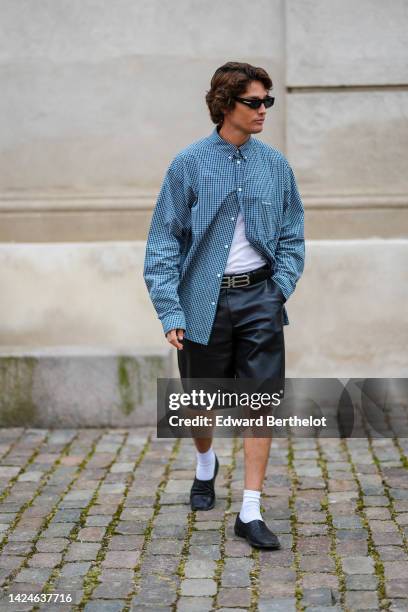 Guest wears black sunglasses, a navy blue and white checkered print pattern shirt, a white t-shirt, a black shiny leather large silver logo buckle...