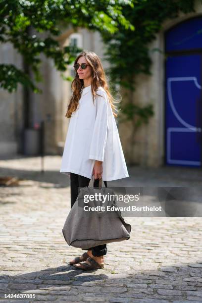 Guest wears brown sunglasses, a white oversized shirt, black pants, a gray fabric large handbag, black belted sandals , outside Aeron, during...