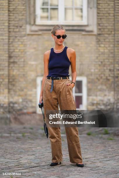 Guest wears black sunglasses, silver earrings, a white pearls necklace, a navy blue tank-top, a black shiny leather belt, brown ribbed velvet wide...
