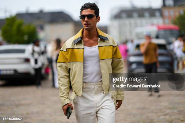 Guest wears black sunglasses, a white tank-top, white latte suit pants, a yellow shiny leather large strap on a pale yellow leather oversized jacket,...